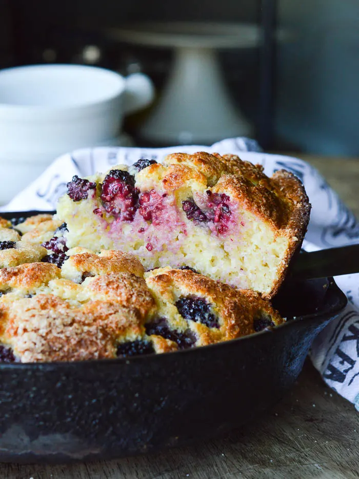 A slice of Blackberry Buttermilk Skillet Cake that's being lifted from the cast iron skillet. the view is from the side so that you can see the warm blackberries that have seeped into the tender crumb of the baked cake. It also shows the fully baked height of about 3 inches tall. 