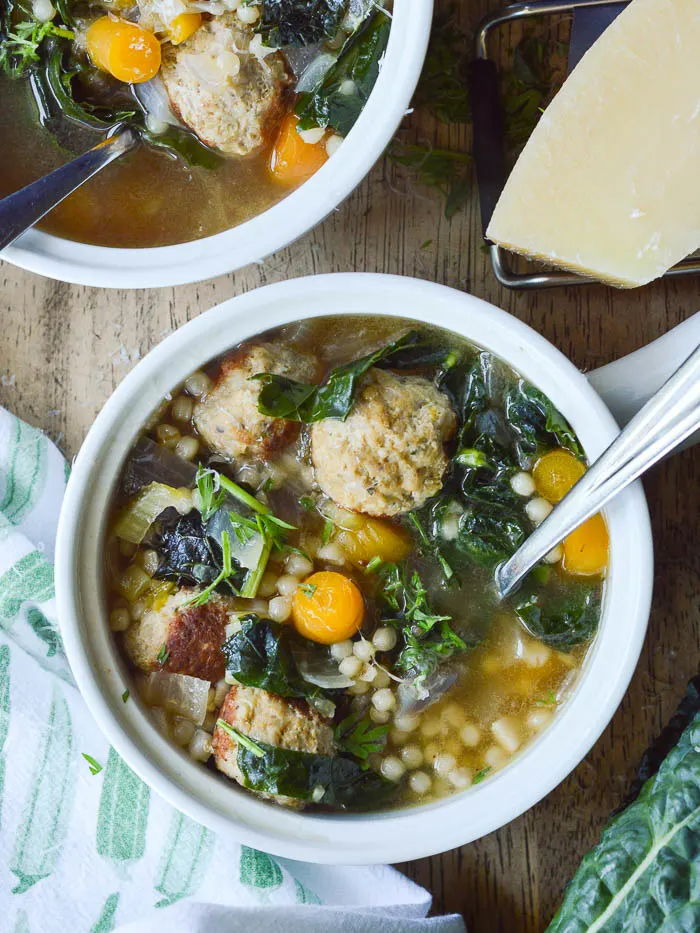 An overhead view of a small crock of Easy Italian Wedding Soup. There are chunks of carrots, torn bits of dark green kale, mini meatballs, israeli couscous, and a rich chicken broth. You can see a small block of Parmesan cheese in the upper righthand corner.