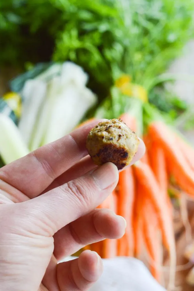 A close up of a bite size meatball held between thumb and forefinger to show the size. There are vegetables needed for soup-making in the background
