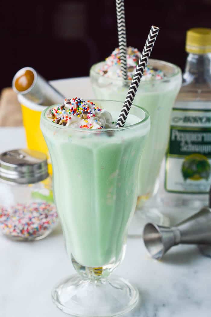Boozy Shamrock Schnapps Shakes in pretty milkshake glasses with paper straws, whipped cream, and rainbow sprinkles