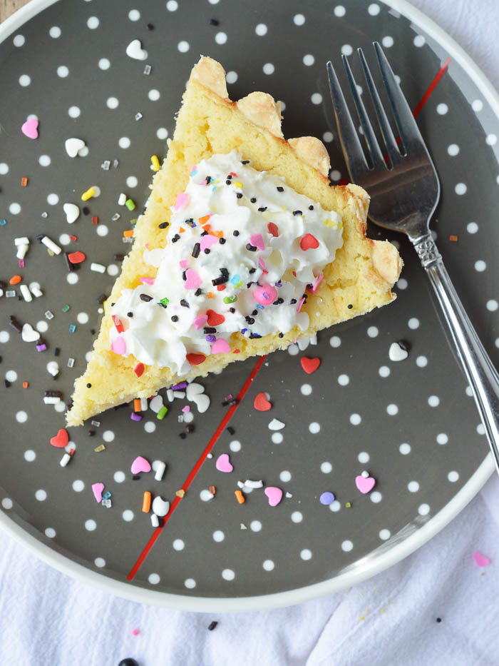 overhead view of Irish Lemon Pudding tart on a gray plate with white polka dots. The slice of tart is topped with a dollop of whipped cream and colorful sprinkles.
