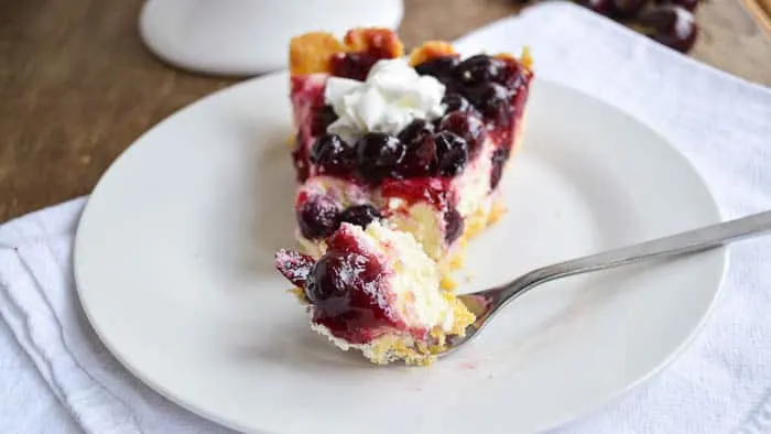 a slice of cheesecake pie topped with cooked cherries and berries, plated with one bite on a fork