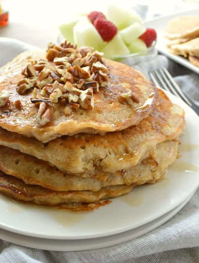 A stack of Banana Oatmeal Pancakes topped with chopped pecans and served with a side of fruit. 
