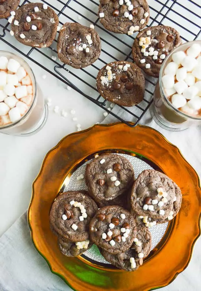 Chocolate chocolate chip cookies studded with tiny mallow bits on a gold-rimmed plate with two big cups of hot chocolate loaded with marshmallows and more cookies on a cooling rack. 