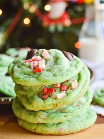 Four green cookies studded with chocolate chips and peppermint chips stacked on a table in front of a Christmas tree