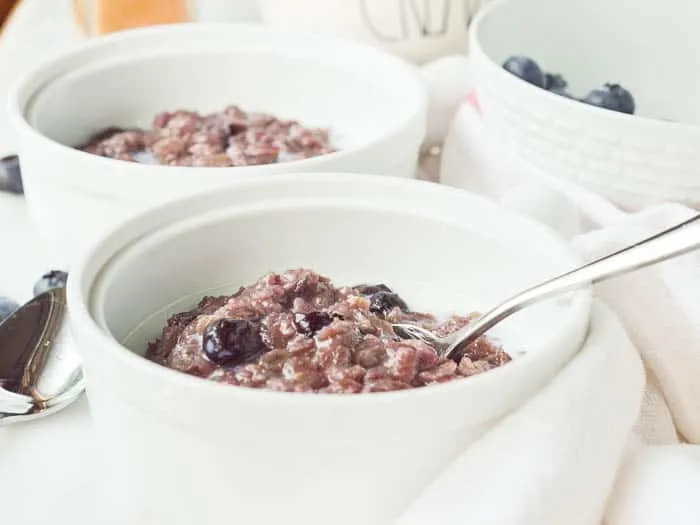Landscape image of Blueberry Cinnamon Oatmeal with a spash of milk and a spoon in the bowl
