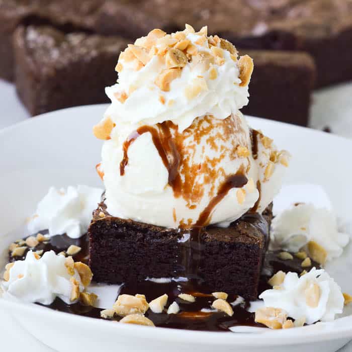 Square cropped image of the best homemade brownie sundae topped with hot fudge, whipped cream, and chopped peanuts