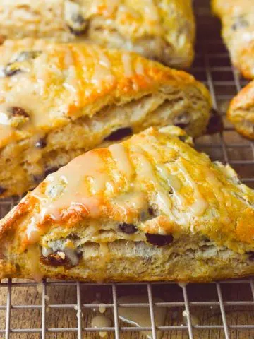 A square cropped close up image of golden brown run raisin scones, glazed and cooling on a cooling rack.