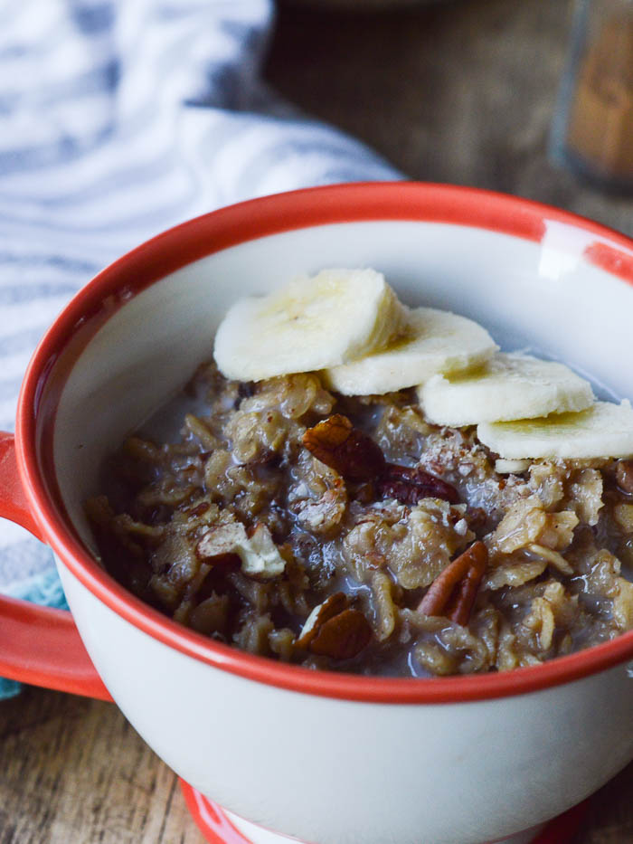 This image of banana cinnamon oatmeal shows cooked old fashioned oats mixed with chopped pecans, ripe bananas, and almond milk in a teacup. Topped with sliced bananas and ready to eat. 