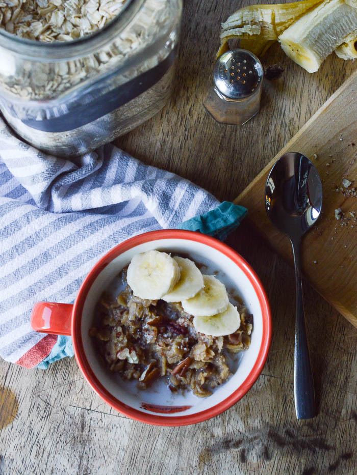 All the makings of banana cinnamon oatmeal - a canister of dry oats, a peeled and sliced banana, a few scattered chopped pecans, and a cinnamon shaker. There's a cup filled with cooked oatmeal ready to eat as well. 