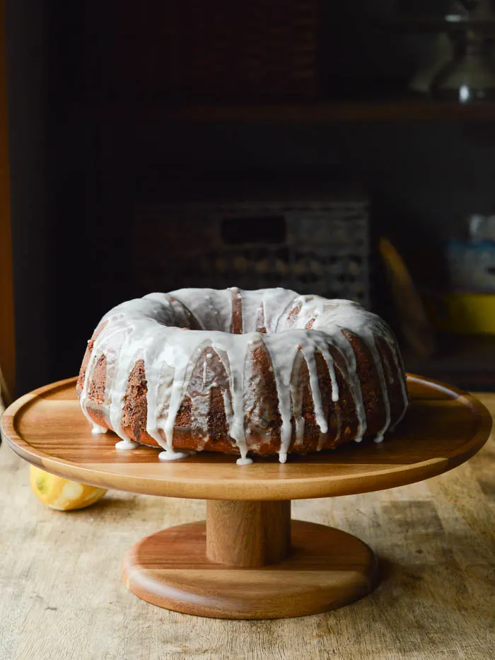 A glazed Olive Oil Bundt Cake on a wooden cake stand with a zested lemon in the background
