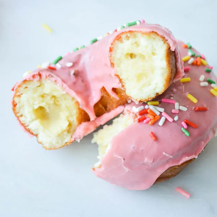 A broken cake doughnut with pink frosting and sprinkles. It's positioned so you can see the perfectly fluffy and tender cake insides and the golden brown cake color on the outside of the donut. 