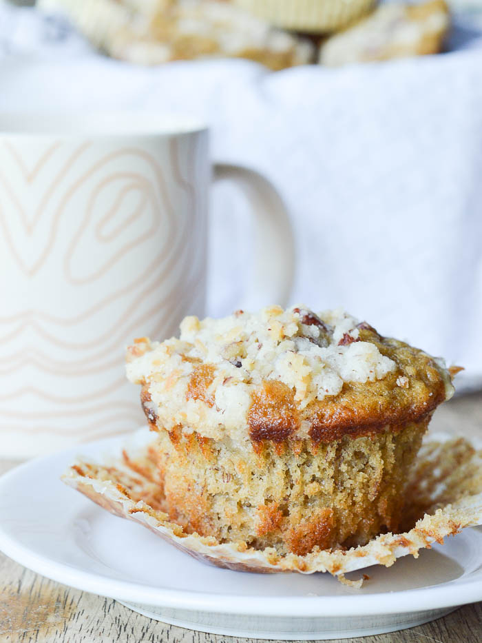 A perfectly golden and fluffy banana muffin with a crumbly pecan streusel topping sitting on a small white plate. The paper liner is peeled away so you can see the flecks of banana and there's a coffee cup in the background. 