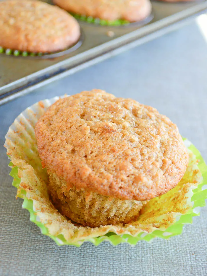 A close up image of a fluffy banana muffin with no topping. It's sitting on the counter with the paper liner peeled back to reveal the soft crumb and you can see the full muffin tin in the background. 