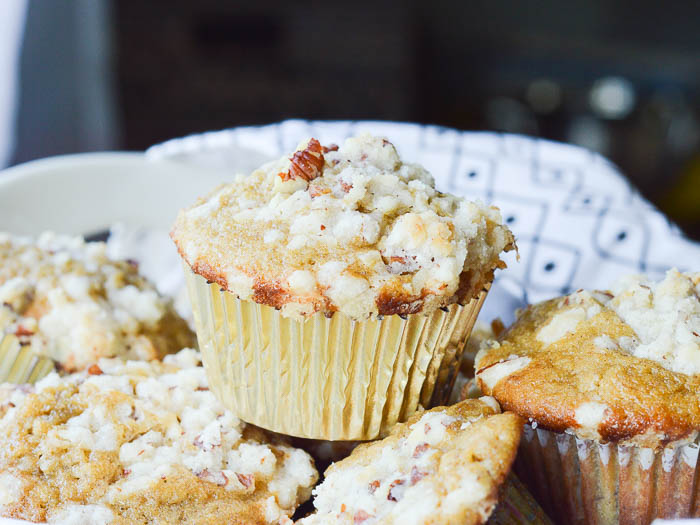 A landscape image of muffins piled in a bowl lined with a black and white tea towel. The focal point of the photo is a perfectly domed muffin in a gold paper liner with a crumbly streusel topping. 