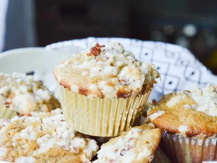 A landscape image of muffins piled in a bowl lined with a black and white tea towel. The focal point of the photo is a perfectly domed muffin in a gold paper liner with a crumbly streusel topping.