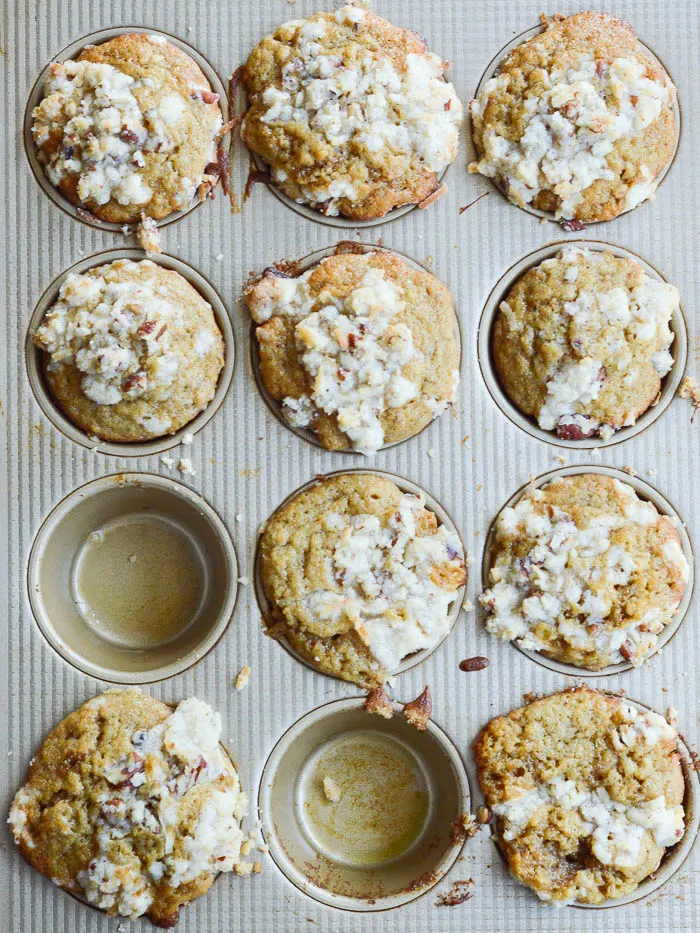 An overhead view of banana muffins in the muffin tin, fresh from the oven. The pan holds 12 and there are two muffins missing, like as if someone needed to eat them warm right away. These banana muffins are topped with streusel and you can see bits of pecans and shredded coconut peeking through the crumbly topping.