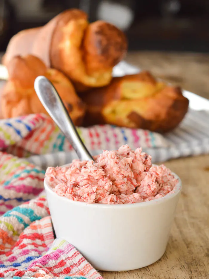 Whipped Strawberry Butter in a white ramekin with a short butter knife ready for serving. There's a pile of warm popovers in the background on a tea cloth.