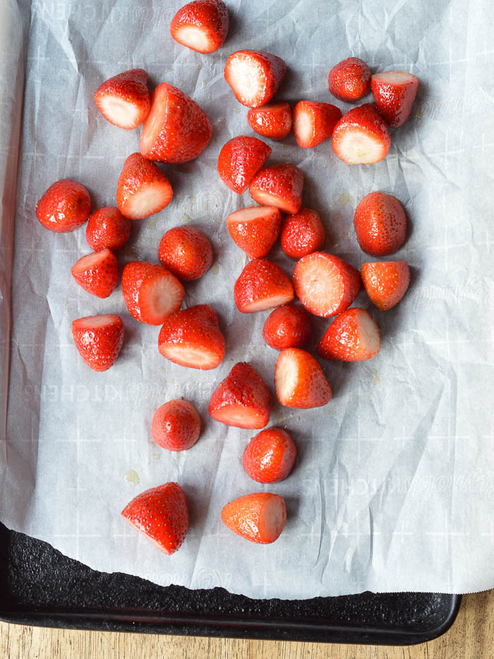 Bright red fresh strawberries that have been hulled, tossed in olive oil, and scattered across a sheet of white parchment paper on a baking sheet. They are ready to be roasted. 