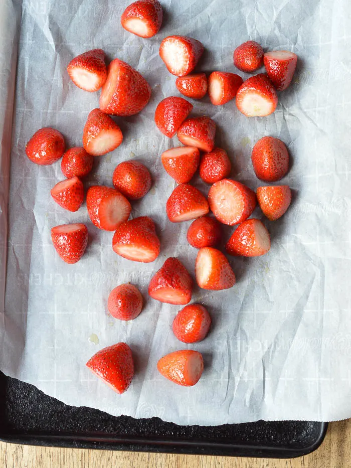 Bright red fresh strawberries that have been hulled, tossed in olive oil, and scattered across a sheet of white parchment paper on a baking sheet. They are ready to be roasted.