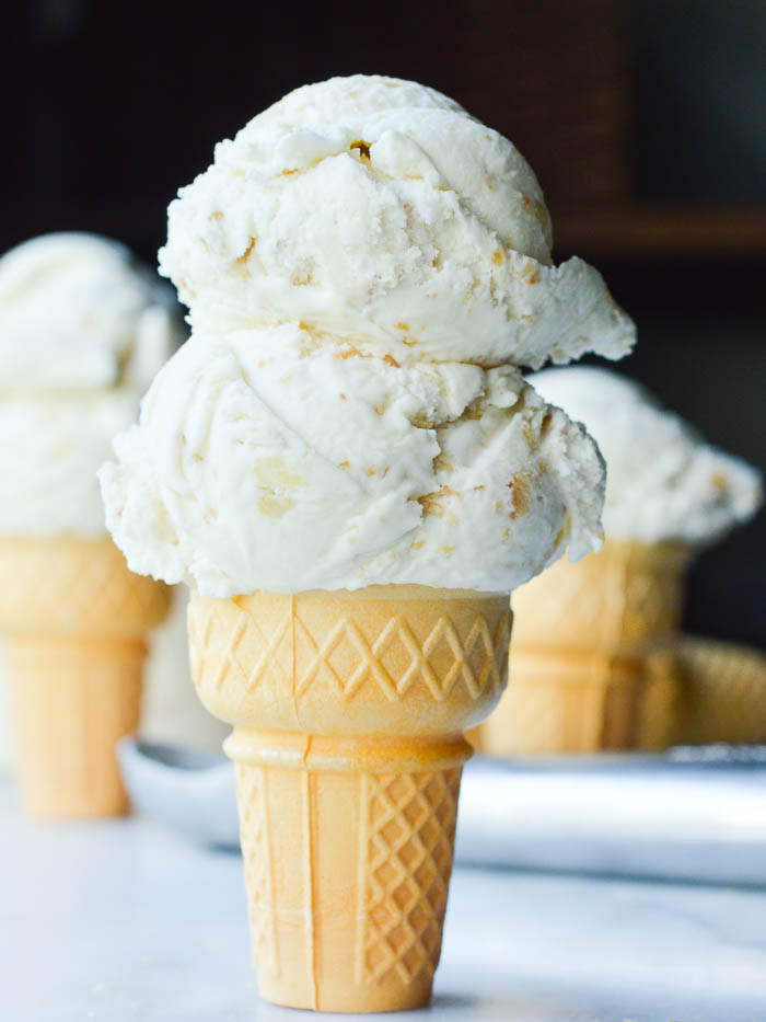 A double scoop of Coconut Macadamia Nut Ice Cream on a sugar cone standing on a white marble pastry board. The ice cream is creamy and studded with nuts. 