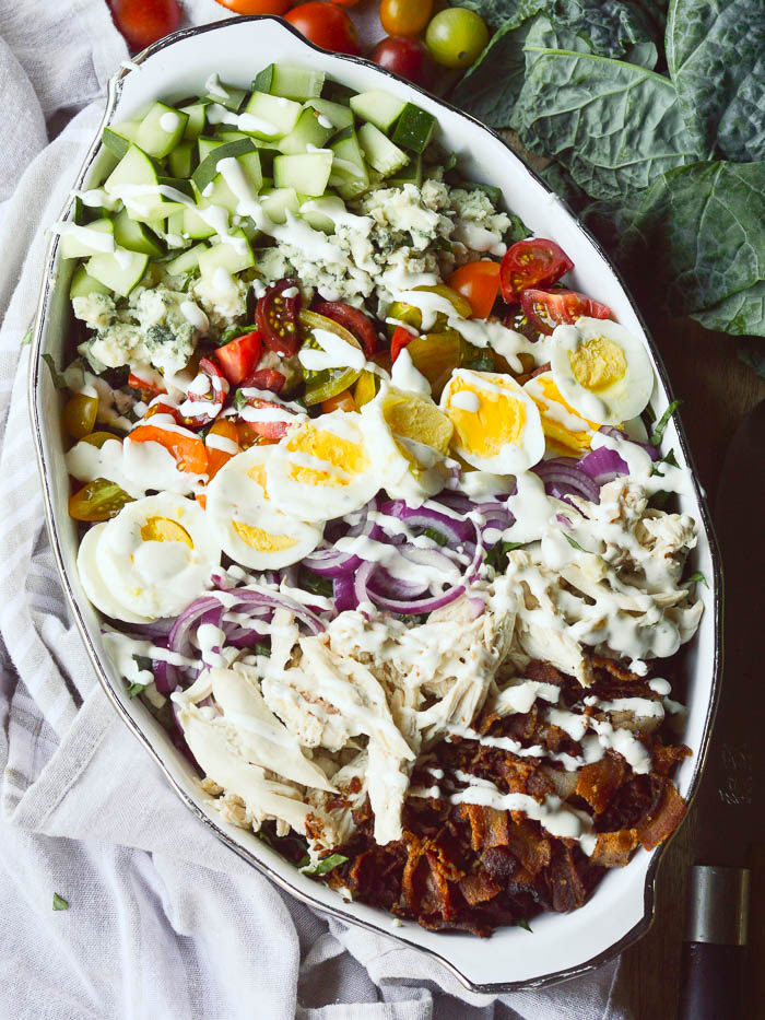 An overhead view of a salad in an oval casserole dish with leaves of dinosaur kale and whole cherry tomatoes peeking in from the background.  Cucumbers, Roquefordt cheese, cherry tomatoes,  hard boiled eggs, sliced onions, shredded chicken, and crumbled bacon are all laid out in rows over a bed of kale and then drizzled with ranch dressing. 
