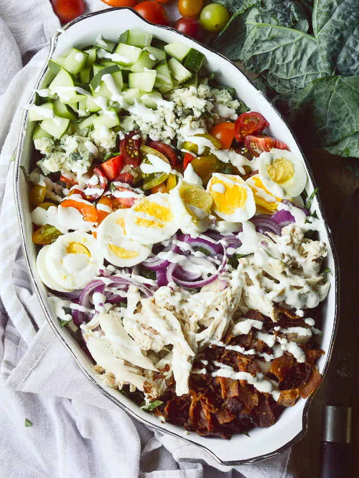 An overhead view of a salad in an oval casserole dish with leaves of dinosaur kale and whole cherry tomatoes peeking in from the background. Cucumbers, Roquefordt cheese, cherry tomatoes, hard boiled eggs, sliced onions, shredded chicken, and crumbled bacon are all laid out in rows over a bed of kale and then drizzled with ranch dressing.