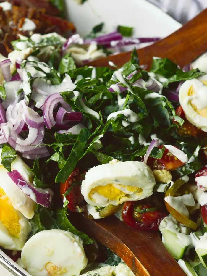 A close up of thinly chopped kale salad drizzled with ranch dressing. There are sliced hard boiled eggs, thinly sliced red onions, chopped cherry tomatoes, bacon, and cucumbers in view. 