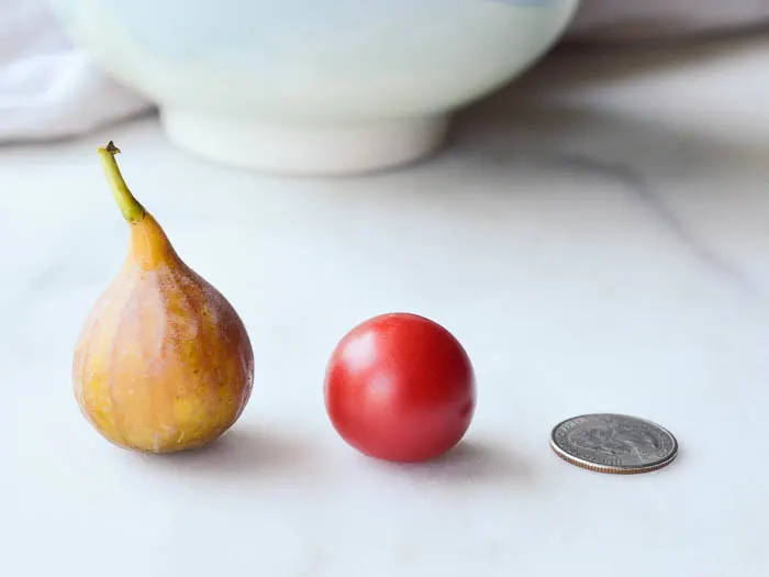 A fresh fig, a cherry tomato, and a quarter all lined up in a row so that you can see the size of the fig to scale. 