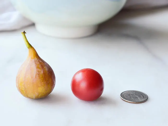 A fresh fig, a cherry tomato, and a quarter all lined up in a row so that you can see the size of the fig to scale.
