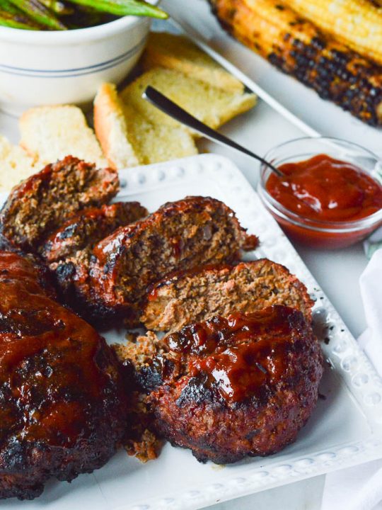 Thick slices of grilled meatloaf stacked on a plate and slathered with spicy ketchup. On the table there's a dish with more ketchup, slices of garlic bread, grilled corn, and grilled okra