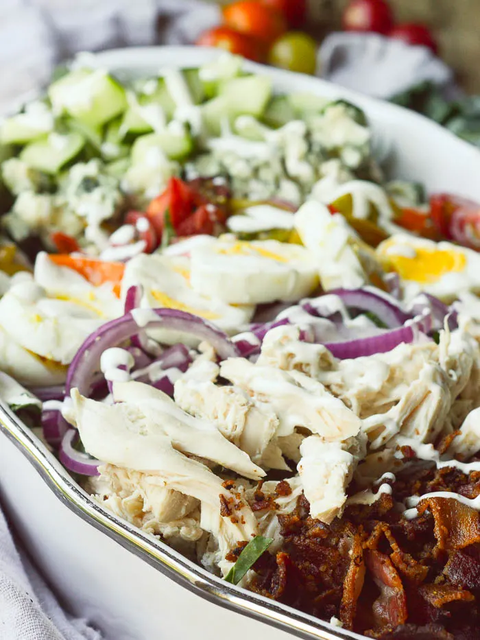 A zoomed in view of shredded rotisserie chicken, sliced red onions, sliced hard boiled eggs, chopped cherry tomatoes and cucumbers, and crumbly Roquefort cheese. Everything is drizzled with ranch dressing. 