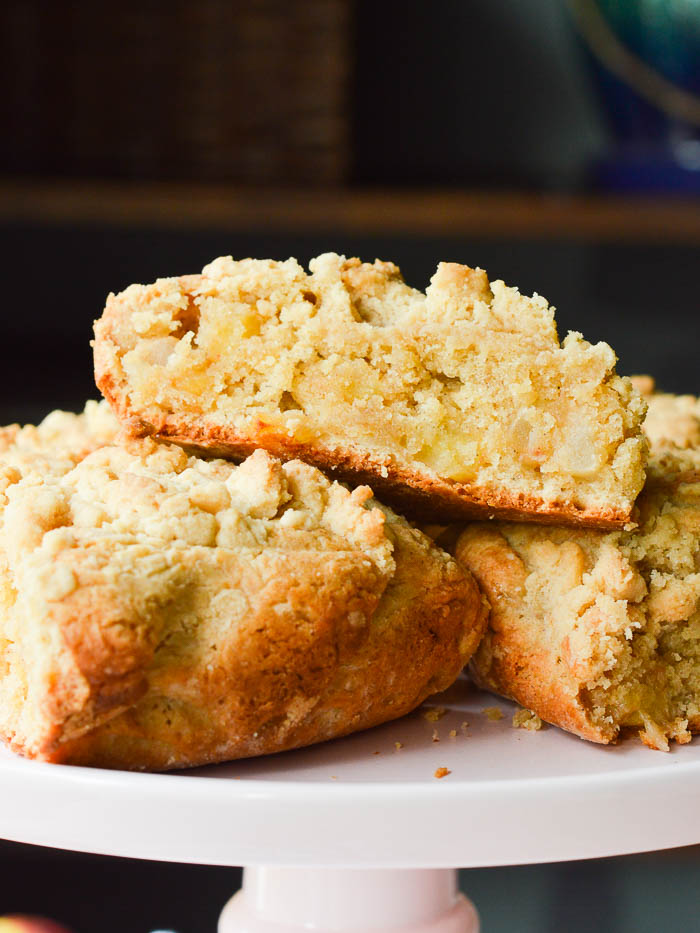 A pile of BIG FAT scones loaded with fresh apples and topped with a buttery streusel. These Apple Pie Scones are sitting atop a cake stand, and you can see the tender cumb of the scones, the crisp baked edges, and a hint of apple pie spice. 