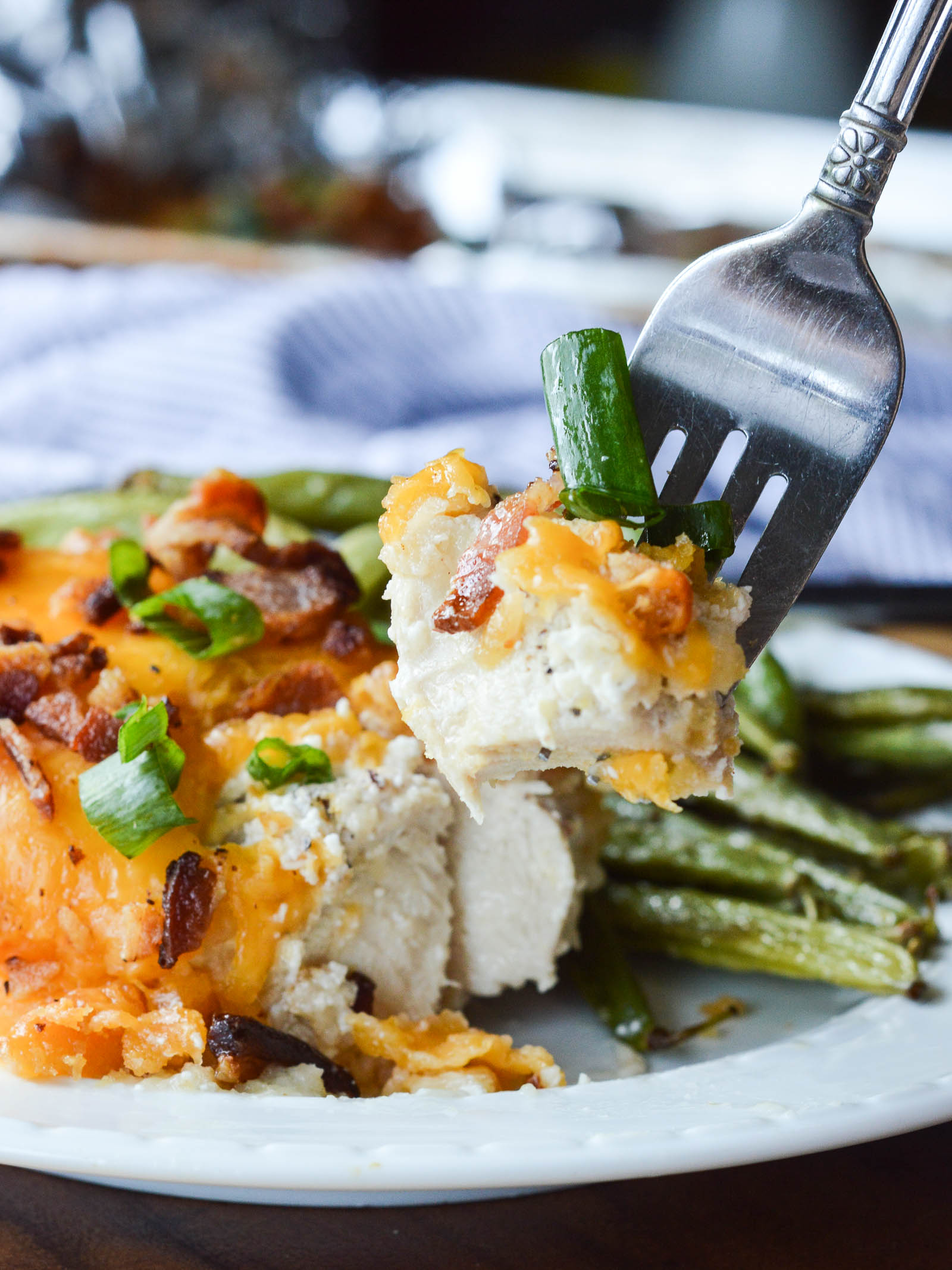 A bite of Smothered Cheesy Sour Cream Chicken on the end of a fork that hovers over the plate. You can see the layers of the chicken dish - seasoned sour cream, melted cheese, crumbled bacon, and chopped green onions. 