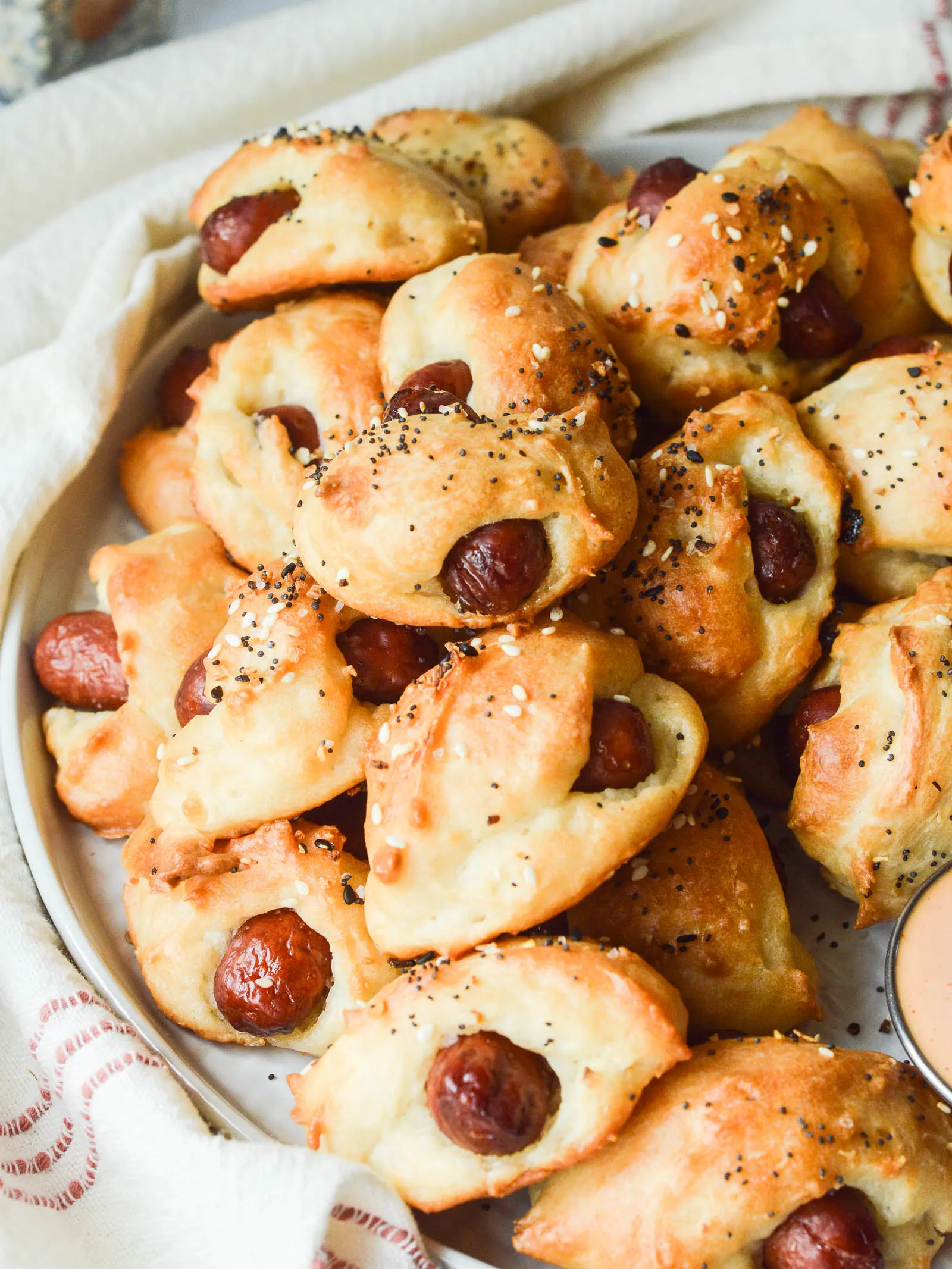 Pigs in a Blanket appetizers that are sprinkled with everything-bagel seasoning, piled onto a plate wrapped with a tea towel.