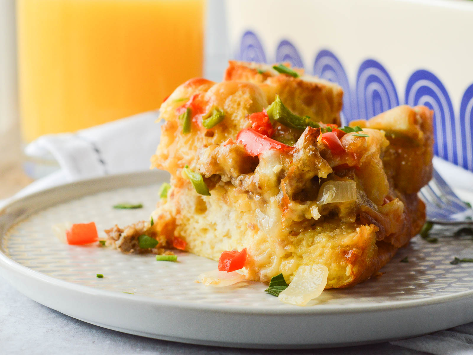 Breakfast Strata with sausage plated with a glass of orange juice in the background. You can see the layers of sausage, peppers, egg and cheese in the baked casserole. 