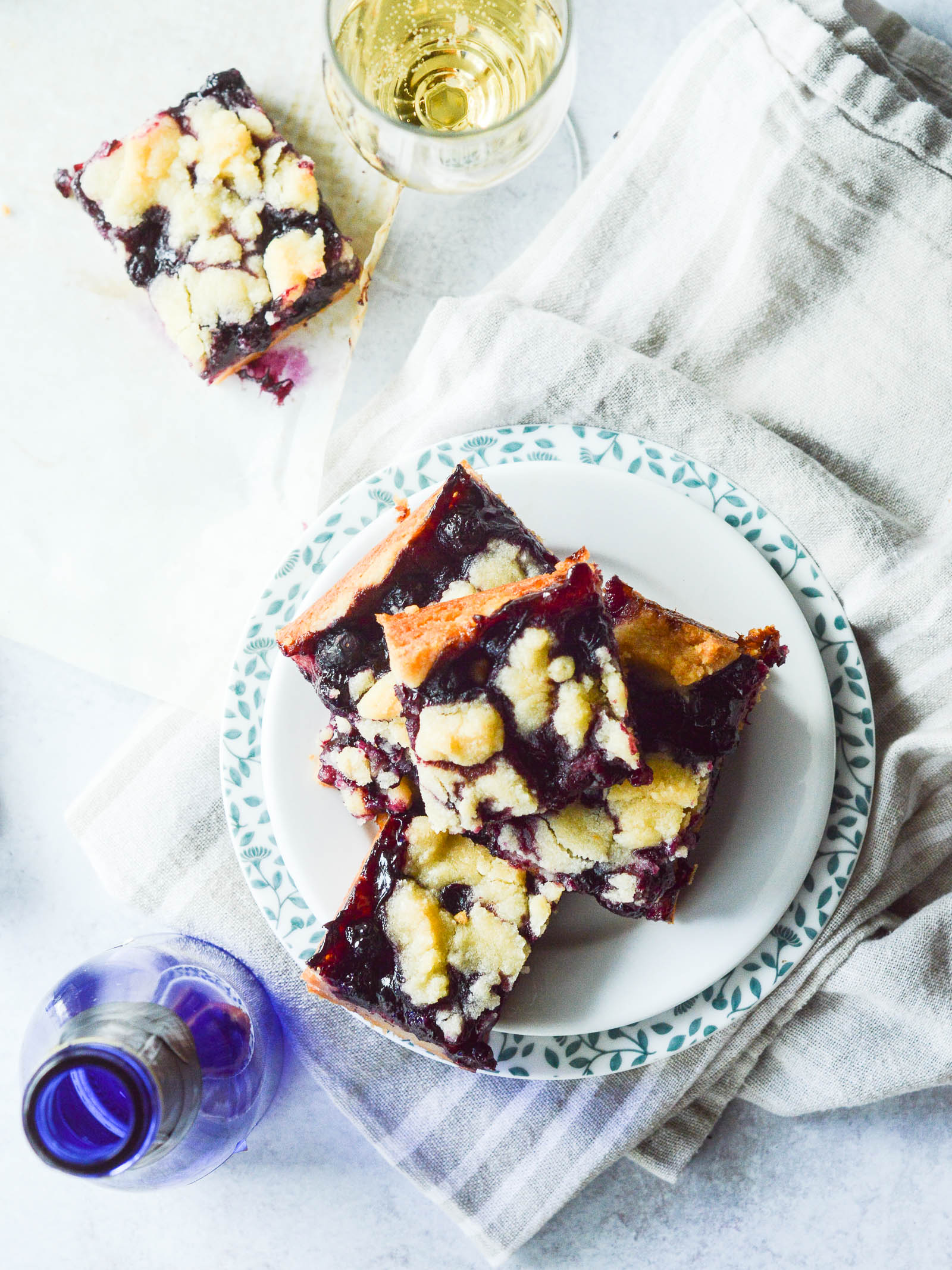 Overhead view of 5 plated Blueberry Crumble Bars on a tea towel with a poured glass of champagne nearby. 