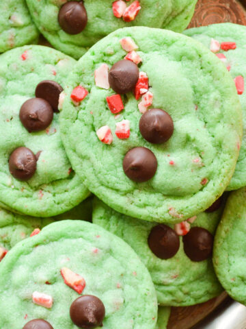 The best Grinch Cookies all piled up on a wooden serving board. they are green and studded with chocolate chips