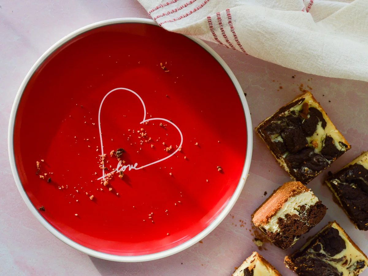 A red plate with a heart on it and the word "love". There area few graham cracker crust crumbs and a board full of cheesecake brownie bars off to the right side. 
