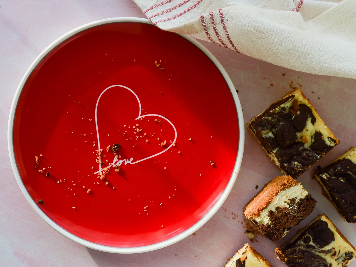 A red plate with a heart on it and the word "love". There area few graham cracker crust crumbs and a board full of cheesecake brownie bars off to the right side.