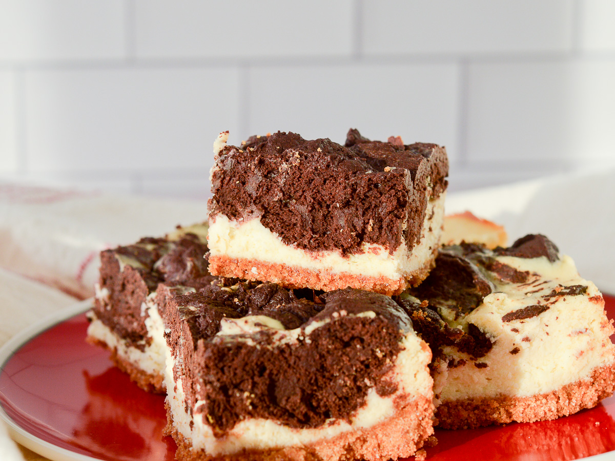 Lancape oriented image of Cheesecake Brownie Bars with a graham cracker crust stacked on a red plate