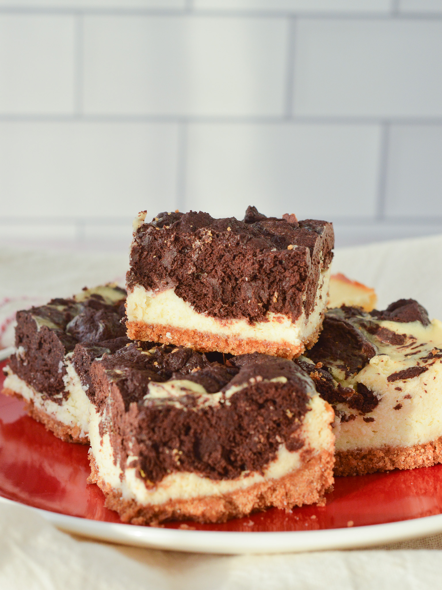 A red plate stacked with cheesecake brownie bars that have a graham cracker crust. 