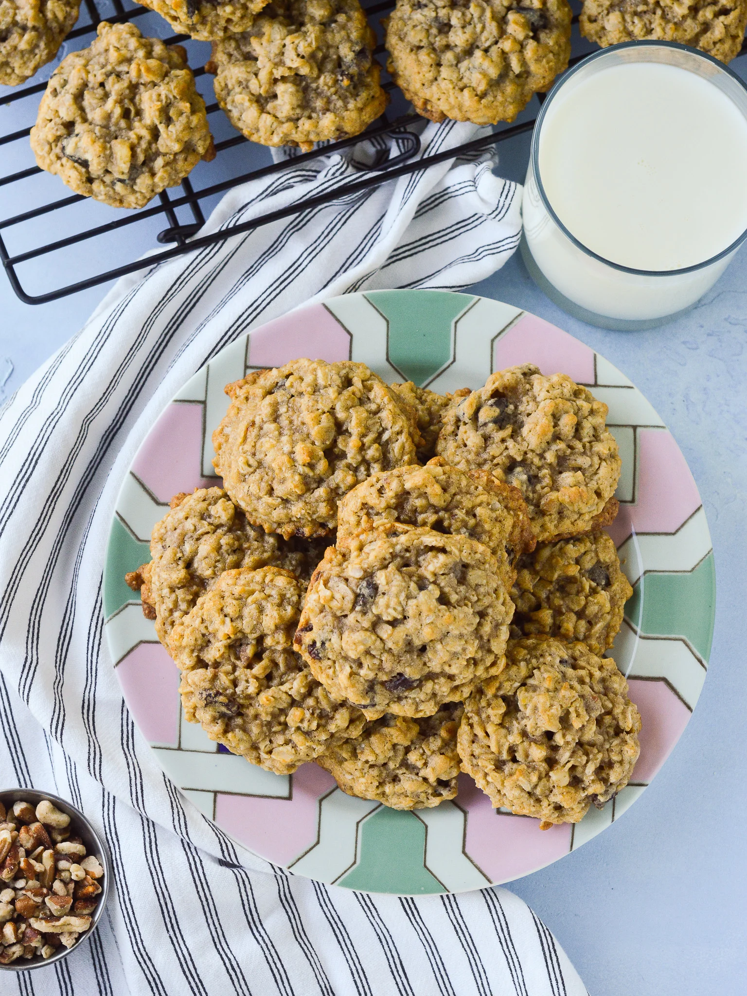 Overhead view of a big plate of Oatmeal Raisin Cookies with a portion of walnuts nearby as well as more cookies cooling on a rack and a glass of milk. 